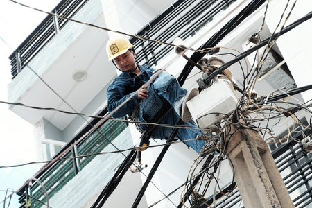 HO CHI MINH CITY, VIET NAM- JULY 12, 2019: Vietnamese electricity worker climb high on electric post to repair electricity network, man work in unsafe service at noon, Vietnamのeditorial素材