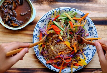 Top view colorful plate of food, vegan mixed vermicelli with vegetables, red, yellow bell pepper, purple cabbage, coriander, cucumber, carrot with mushroom sauce, vegetarian dish on wooden backgroundの写真素材