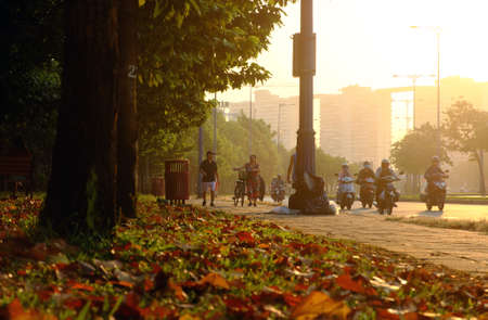 HO CHI MINH CITY, VIET NAM- JAN 15, 2020: Wonderful scene at early morning in park, with leaves from tree fall on grass field, people moving on street in warm sunlight ray, Vietnamのeditorial素材