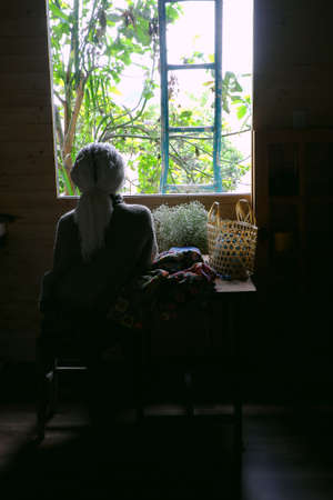 Old woman sitting near window with handmade product, yarn colorful blanket by crochet from yarn with old style, window light make shadow on wooden table of classic styleの写真素材