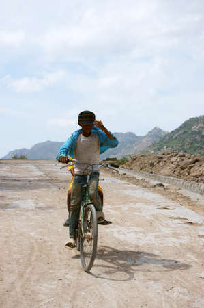 LAM DONG, VIET NAM- FEB 4, 2013: Boy ride a bike go down slope on path of deserted countryside, two Vietnamese children enjoy with outdoor activity under sun, dirty clothing, wear hat, tanned skinの写真素材