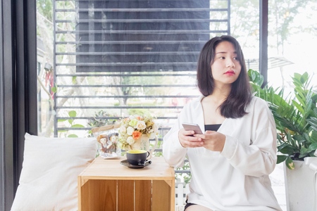 An Asian girl in white coat sitting and playing mobile phone in the glass room of vintage cafe in Bangkok, Thailand January 3,2017のeditorial素材
