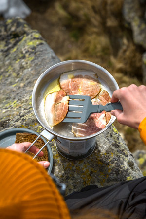 A woman's hand flips a piece of ham on a frying pan to prepare food on a camping trip in the mountainsの写真素材