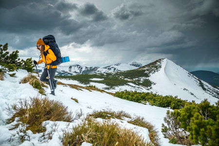 Young woman hiking with hiking pole on snowy mountain against cloudy sky. Active young woman in orange wind jacket carrying backpack hiking in mountainsの写真素材