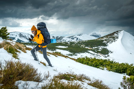Young woman hiking with hiking pole on snowy mountain against cloudy sky. Active young woman in orange wind jacket carrying backpack hiking in mountainsの写真素材