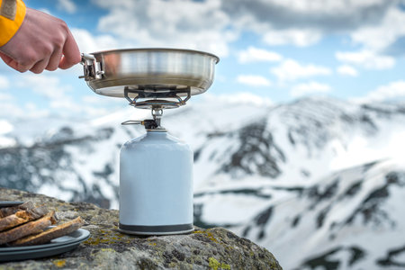 A woman's hand holds camping fry pan on a tourist gas stove to prepare food on a camping trip in the mountainsの写真素材