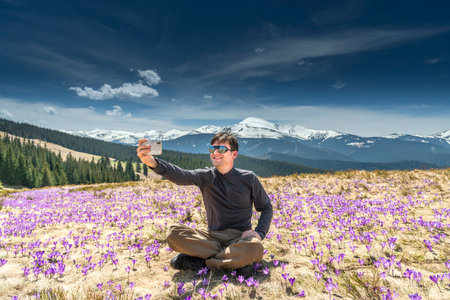 Young hiker man taking selfie while sitting on mountainside full of purple crocuses during summer hikeの写真素材
