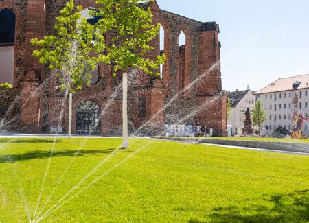 Old red brick church. Partially destroyed walls. Young lawn in front of the entrance. Watering the lawn. Gardener's work. Historical building.の写真素材