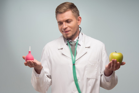 Half-length portrait of very attractive young doctor, wearing white uniform, looking at the enema in her right hand  Isolated on the white の写真素材