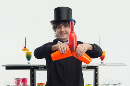 Half-length portrait of handsome smiling bartender wearing nice black topper and white bow-tie showing us different tricks with orange bottles and glassesの写真素材
