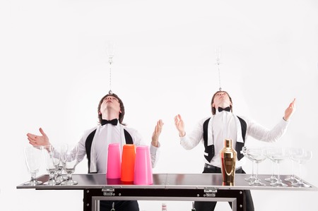 Two handsome barmen standing near the bar counter with different glasses and shaker on it holding on their head bar spoon with empty glass on the topの写真素材