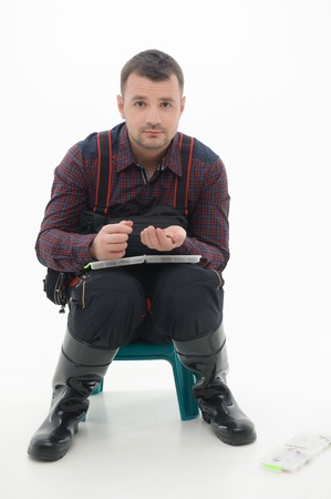 Handsome man wearing nice colorful shirt and black waders sitting on the little table and holding in his hands fishing hooks  Isolated on white backgroundの写真素材