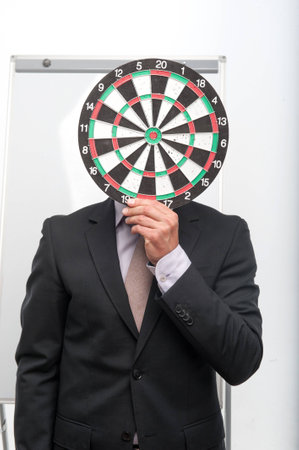 Half-length portrait of man wearing nice suit closing his face with the target for dartsの写真素材