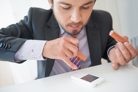 Attractive young businessman taking pleasure in very delicious sausage and in the same time dialing the numberの写真素材