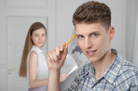 Selected focus on handsome young smiling boy holding pen in his hand and thinking about wonderful scientific project he invented  His project partner on backgroundの写真素材