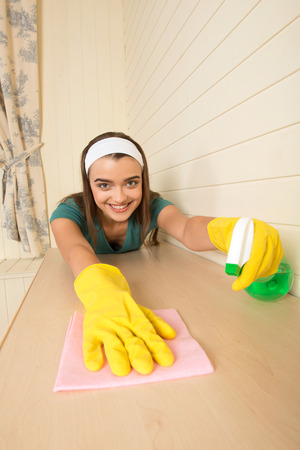 Lovely dark-haired housemaid with the white fillet wearing yellow gloves wiping cream-colored table in the kitchenの写真素材