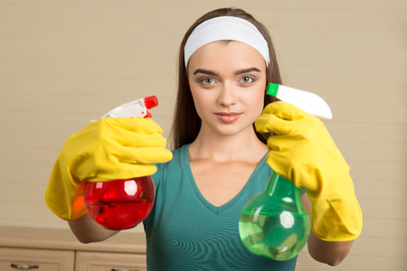 Half-length portrait of beautiful young housemaid wearing rubber yellow gloves showing us red water sprayer in one hand and green in anotherの写真素材
