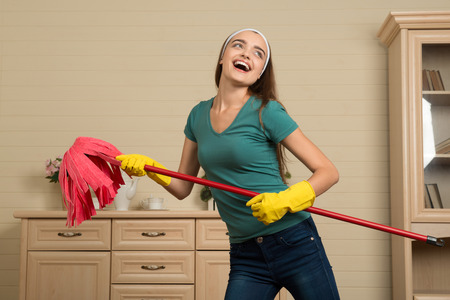 Funny smiling dark-haired housemaid wearing nice shirt and jeans with the mop imagining she is famous artisteの写真素材