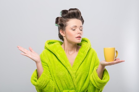 Half-length portrait of young pleasant housewife wearing curlers and green wrapper holding yellow cup of tea and relaxing  Isolated on grey backgroundの写真素材