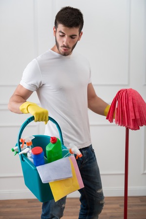 Half-length portrait of young dark-haired janitor wearing white shirt blue torn jeans and yellow rubber gloves standing with the red mop in one hand and pail with cleansers in another looking at somethingの写真素材