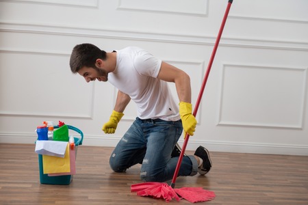 Full-length portrait of very angry dark-haired janitor wearing white shirt blue torn jeans and yellow rubber gloves sitting on the floor with the red mop and pail with cleansers he hates his jobの写真素材