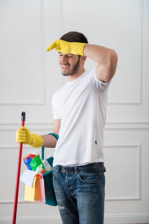 Full-length portrait of very tired dark-haired janitor wearing white shirt blue torn jeans and yellow rubber gloves standing with the red mop and pail with cleansers wiping her head with her handの写真素材