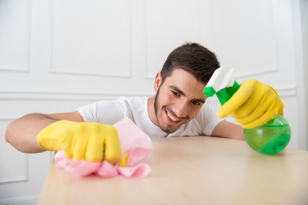 Half-length portrait of young smiling dark-haired janitor wearing white shirt and yellow rubber gloves dusting the tableの写真素材
