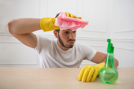 Half-length portrait of young tired dark-haired janitor wearing white shirt and yellow rubber gloves sitting at the table holding pink duster in the hand groaningの写真素材