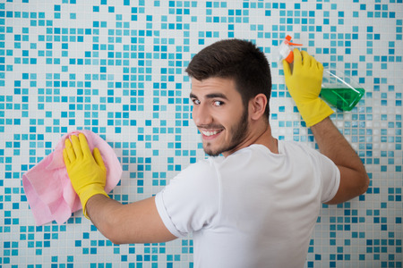 Half-length portrait of young smiling dark-haired janitor wearing white shirt and yellow rubber gloves standing back in the bathroom and cleaning it with the rag and detersiveの写真素材