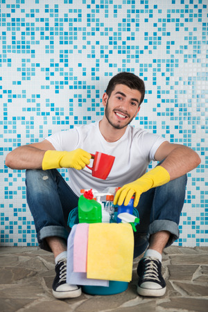 Young smiling dark-haired janitor wearing white shirt blue jeans and yellow rubber gloves sitting in the bathroom with pail of cleaners and cup of coffee satisfied with his workの写真素材