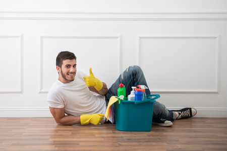 Young dark-haired janitor wearing white shirt blue jeans and yellow rubber gloves lying on the floor near pail of cleaners relaxingの写真素材