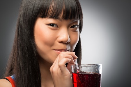 Pretty young smiling dark-haired Asian woman drinking her favorite cherry juice through the straw  Isolated on black backgroundの写真素材