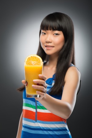 Half-length portrait of pretty young smiling dark-haired Asian woman showing us her orange juice with half of orange in it  Isolated on black backgroundの写真素材