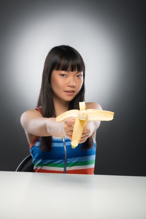 Half-length portrait of pretty young sexy dark-haired Asian woman sitting at the table and pointing banana at us  Isolated on black backgroundの写真素材