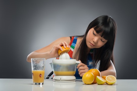 Half-length portrait of pretty young dark-haired Asian woman sitting at the table squeezing juice from oranges and lemons using juicer very diligently  Isolated on black backgroundの写真素材