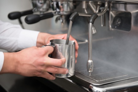 Hands of professional barista warming the milk in the special metal glass in the coffee machine  の写真素材