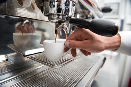 Hands of professional barista holding two white cups on the grating of coffee machine looking how coffee pouring into themの写真素材