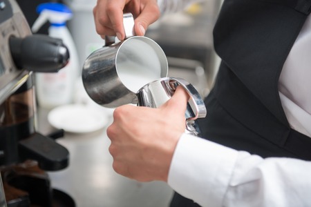 Selected focus on the professional barista wearing white shirt and black apron pouring milk from big metal jar to the small one  Coffee machine on backgroundの写真素材