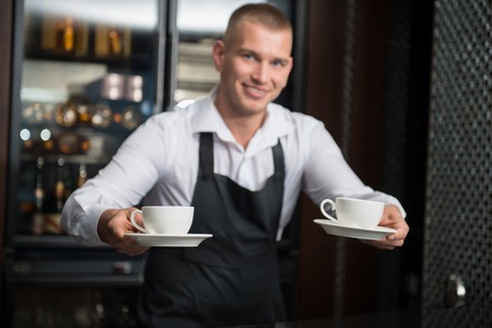 Selective focus on two white cups in the hands of handsome young smiling barista wearing white shirt and black apron suggested us to taste his coffee on backgroundの写真素材
