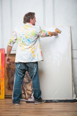 Full-length portrait of fair-haired painter wearing painted white shirt and blue torn jeans standing back to us among his pictures bleaching the linenの写真素材