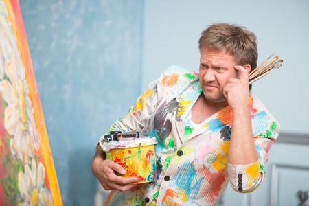 Half-length portrait of fair-haired anxious painter wearing white painted jacket standing with brushes and tin of paints looking at his picture does not know what to addの写真素材
