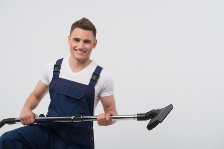Half-length portrait of young dark-haired smiling janitor wearing white T-shirt and overalls standing with the big vacuum cleaner showing it us  Isolated on white backgroundの写真素材