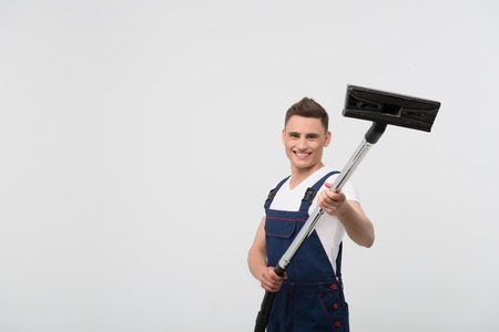 Half-length portrait of young dark-haired smiling janitor wearing white T-shirt and overalls standing with the big vacuum cleaner showing it us  Isolated on white backgroundの写真素材