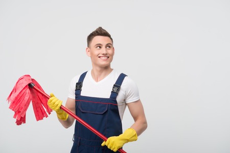 Half-length portrait of young smiling dark-haired janitor wearing white T-shirt overalls and yellow rubber gloves standing with the red mop  Isolated on white background の写真素材
