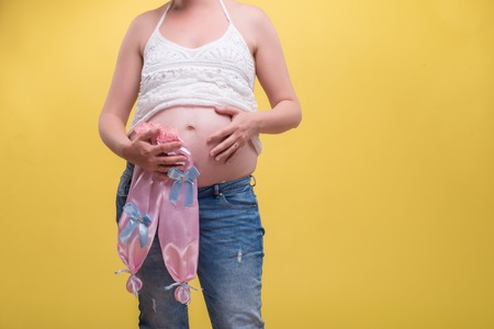 Half-length portrait of young pretty dark-haired smiling pregnant woman wearing white T-shirt and jeans standing with small pink pants for baby girl touching her belly. Isolated on yellow backgroundの写真素材