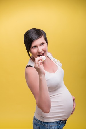 Half-length portrait of young dark-haired funny pregnant woman wearing white T-shirt and jeans standing aside holding a pacifier in her mouth. Isolated on yellow backgroundの写真素材