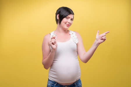 Half-length portrait of young dark-haired smiling pregnant woman wearing white T-shirt and jeans standing holding a pacifier and pointing at someone. Isolated on yellow backgroundの写真素材