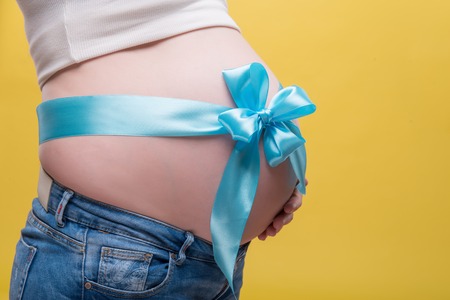 Half-length portrait of pregnant woman wearing white T-shirt and jeans standing aside with blue band on her belly dreaming about her baby. Isolated on yellow backgroundの写真素材