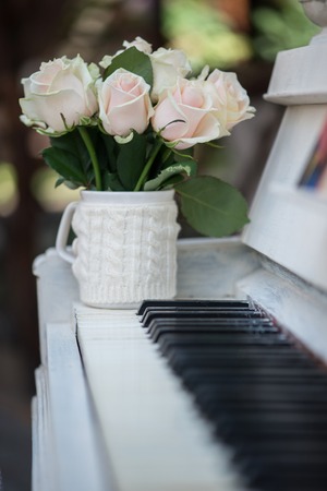Selective focus on beautiful little bouquet of white roses standing in the nice decorated cup on the white opened piano on backgroundの写真素材