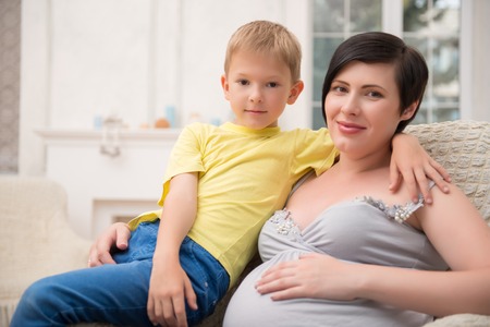 Selected focus on dark-haired smiling pregnant woman wearing nice shirt and black pants sitting in...